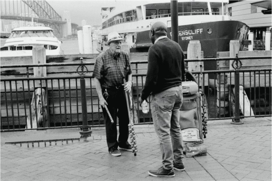 Sydney: two men talking at the harbour railing, a ferry and the Harbour Bridge behind them.