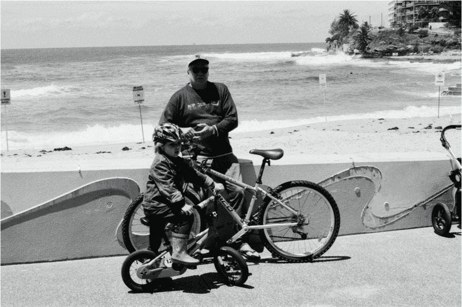 Sydney: a man resting on his bicycle beside a child on a small bike, the surf behind them.